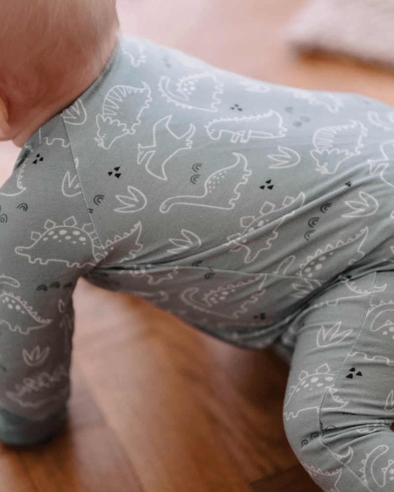 A baby crawling on a wooden floor wearing a sage green long-sleeve one-piece jumpsuit with a white dinosaur pattern.