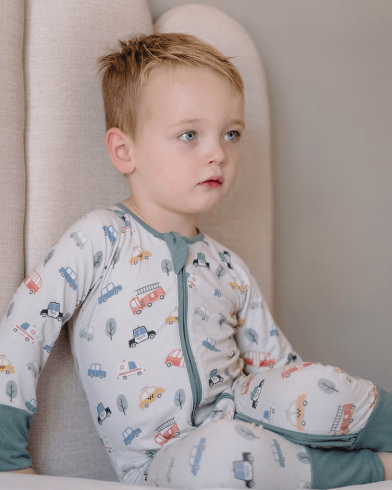A young child wearing a long-sleeve footless pajama top and matching pants with a cute car and truck pattern, sitting against a neutral beige upholstered chair.
