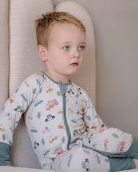 A young child wearing a long-sleeve footless pajama top and matching pants with a cute car and truck pattern, sitting against a neutral beige upholstered chair.