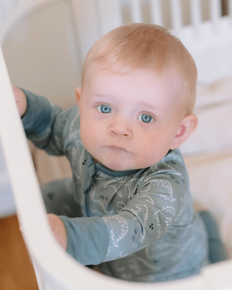 A baby with light hair and striking blue eyes wearing a slate blue long-sleeve onesie with a white dinosaur pattern, sitting inside a white crib.