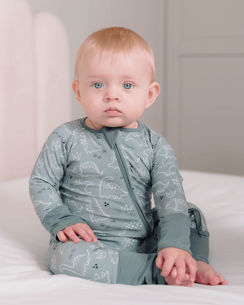 A baby with light hair and blue eyes sits on a white bed wearing a sage green long-sleeve pajama with a white dinosaur pattern.