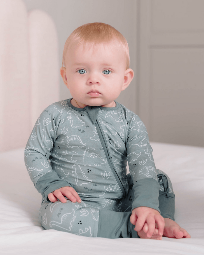 A baby with light hair and blue eyes sits on a white bed wearing a sage green long-sleeve pajama with a white dinosaur pattern.