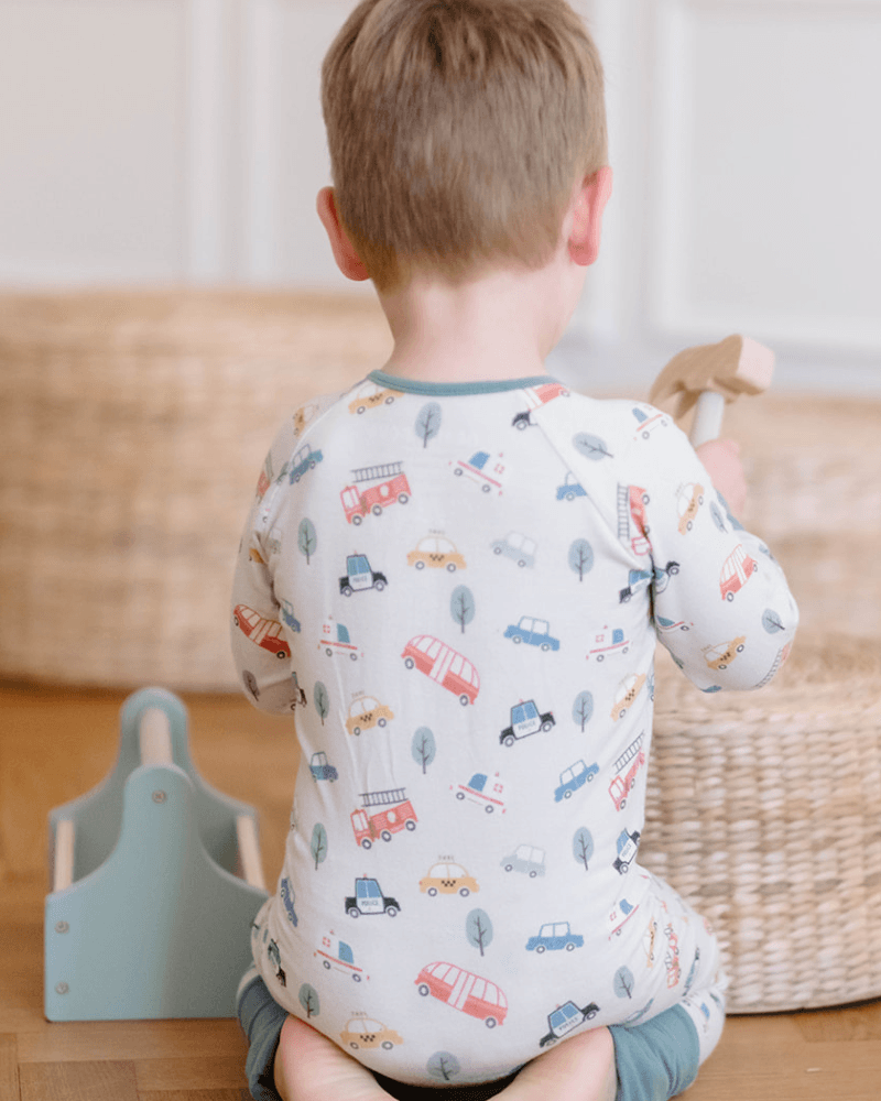 A toddler seen from behind wearing a white long-sleeve pajama with a colorful car and tree print, sitting on a wooden floor in a nursery setting.