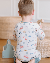 A toddler seen from behind wearing a white long-sleeve pajama with a colorful car and tree print, sitting on a wooden floor in a nursery setting.