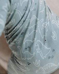 Close-up of a soft fabric item, likely a blanket or pillow, in a dusty sage green color with a white outline print of various dinosaurs.