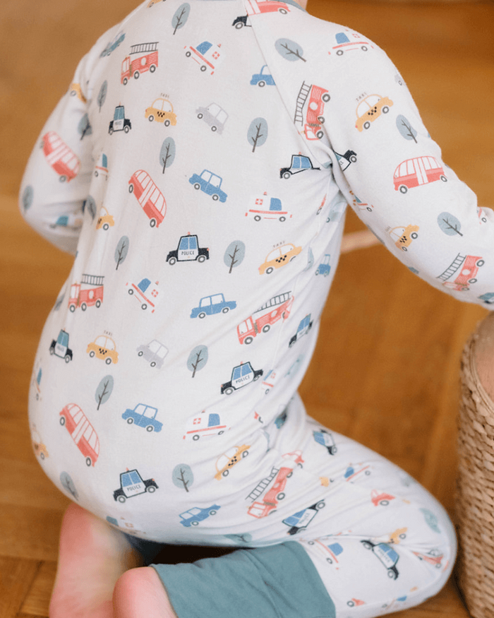 A baby wearing a long-sleeve white one-piece pajama with a colorful cartoon car and truck pattern, kneeling on a wooden floor.
