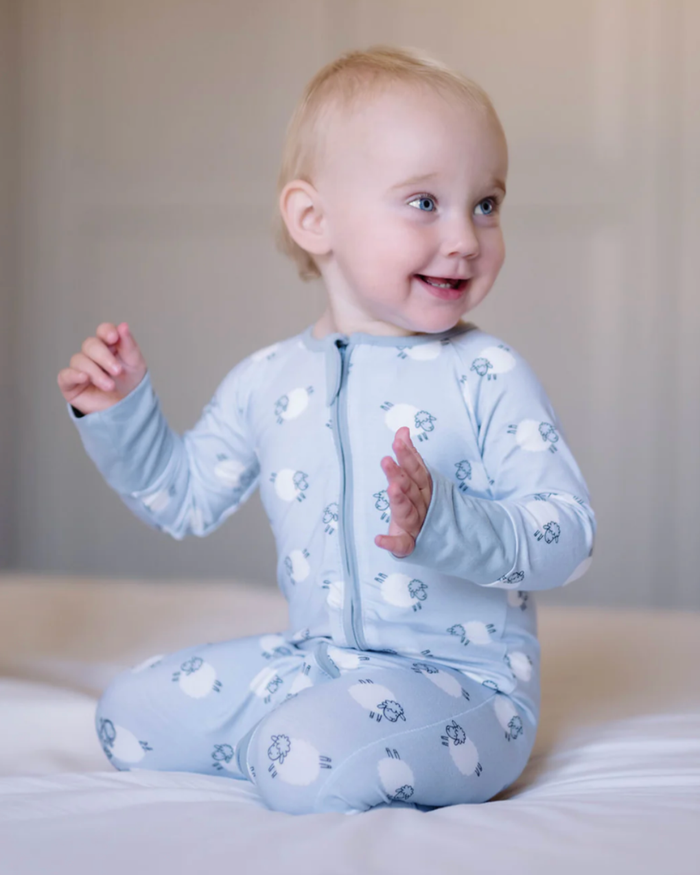 A baby sitting on a white bed wearing a light blue long-sleeve zip-up sleeper with a sheep pattern.