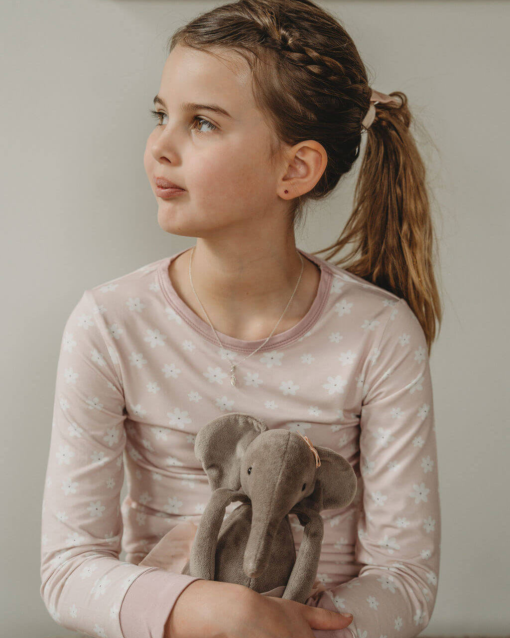 A young girl with braided hair in a ponytail sits on a carpeted floor, wearing a pale pink long-sleeve pajama top with a white floral pattern and holding a gray plush elephant toy.