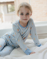 A young girl with blonde hair sits on a white bed wearing a light blue long-sleeve pajama set with a small orange and yellow pattern. The background shows a softly blurred window and brick wall.
