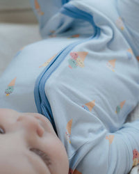 A baby lying on a white blanket wearing a light blue zip-up sleeper with a small pastel ice cream cone pattern.