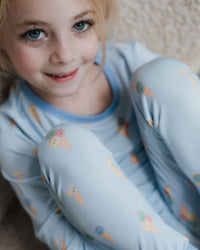 A young girl with blonde hair and blue eyes wearing light blue pajamas with a colorful ice cream cone pattern, sitting on a beige carpet.