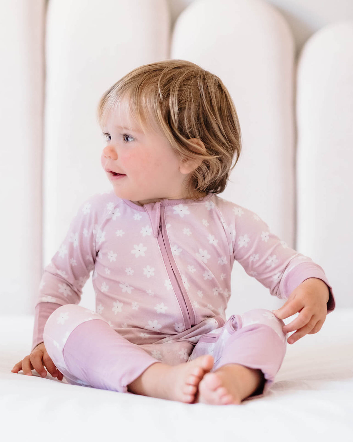 Child wearing a pink star-patterned onesie sitting on a white bed.