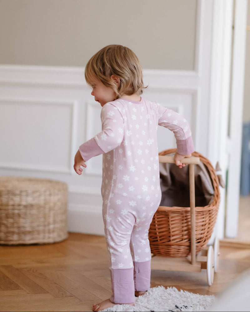 A toddler stands barefoot in a room, wearing a light pink long-sleeve pajama with white star prints and matching pink leg warmers. The child is facing away from the camera, looking down, with a wicker basket and wooden floor in the background.