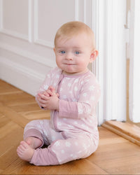 A baby sitting on a wooden floor wearing a light pink long-sleeve footless sleeper with a white snowflake pattern.