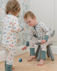 Two children in matching pajamas playing on a stool in a room with floral wallpaper.