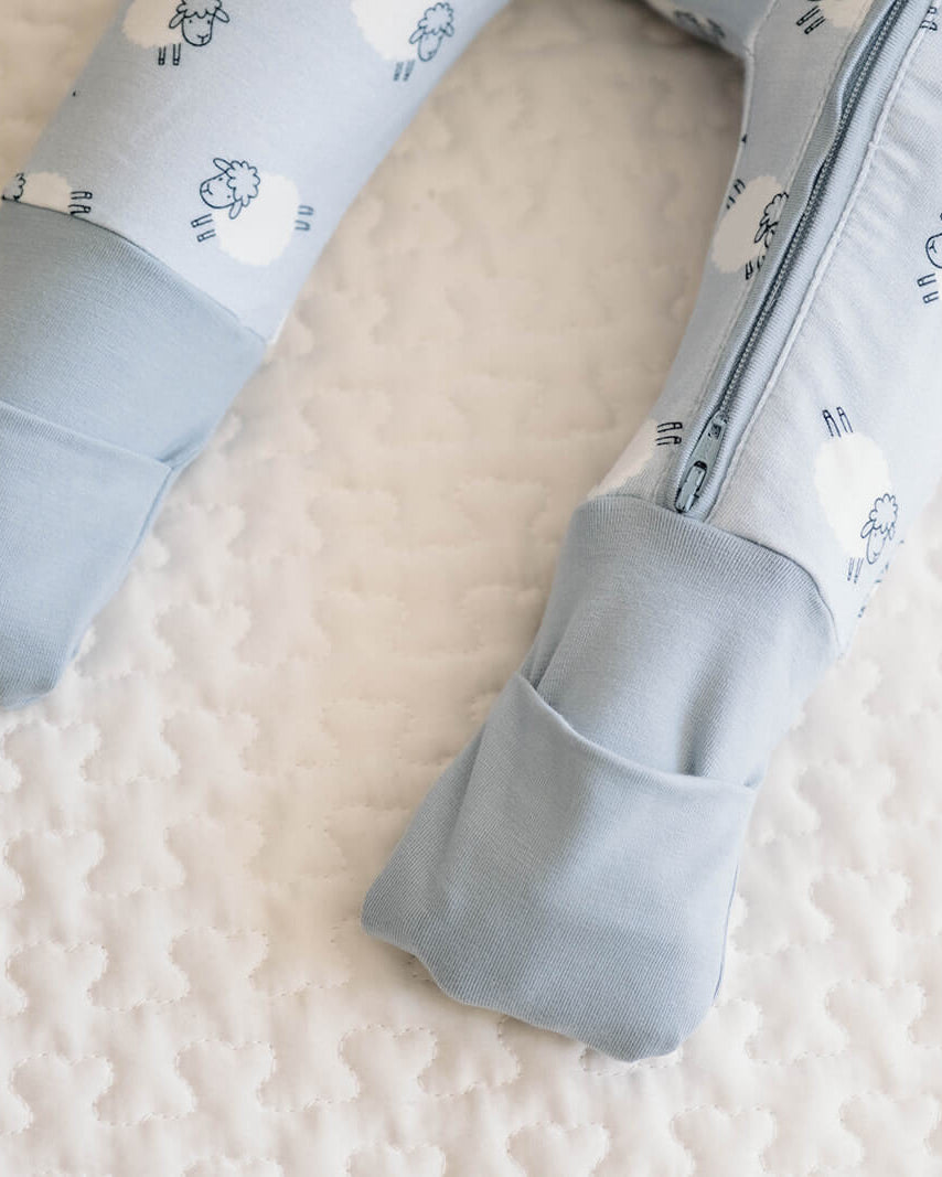 Close-up of a baby's legs wearing light blue footed pajamas with a white cloud pattern, lying on a textured white quilted mattress.