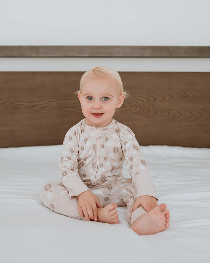 A baby sitting on a white bed with a wooden headboard, wearing a light beige patterned long-sleeve romper.