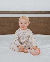 A baby sitting on a white bed with a wooden headboard, wearing a light beige patterned long-sleeve romper.