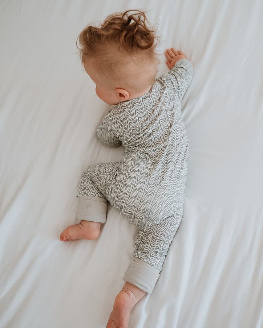 A baby lying on their side on white bedding, wearing a light gray and white patterned long-sleeve footless onesie.