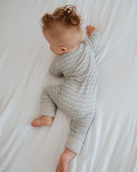 A baby lying on their side on white bedding, wearing a light gray and white patterned long-sleeve footless onesie.