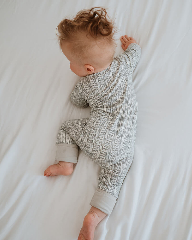 A baby lying on their side on white bedding, wearing a light gray and white patterned long-sleeve footless onesie.
