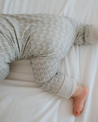 A baby wearing a gray and white textured herringbone patterned long-sleeve romper, lying on white sheets.