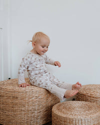 A toddler sits barefoot on a round woven seagrass ottoman in a bright room, wearing a light beige long-sleeve pajama top and matching pants with a small repeating pattern.
