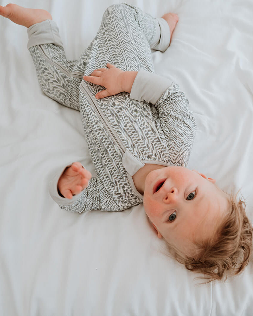 A baby lying on white bedding, wearing a light gray patterned long-sleeve footed sleeper.