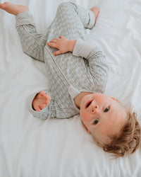 A baby lying on white bedding, wearing a light gray patterned long-sleeve footed sleeper.