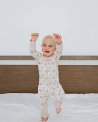 A baby wearing a light beige patterned long-sleeve pajama set stands on a white bed with a dark wood headboard, holding up both fists in a playful pose against a white wall.