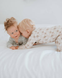 Two toddlers lying on a white bed, wearing matching beige and white patterned pajamas. One toddler is looking directly at the camera, while the other leans in close.