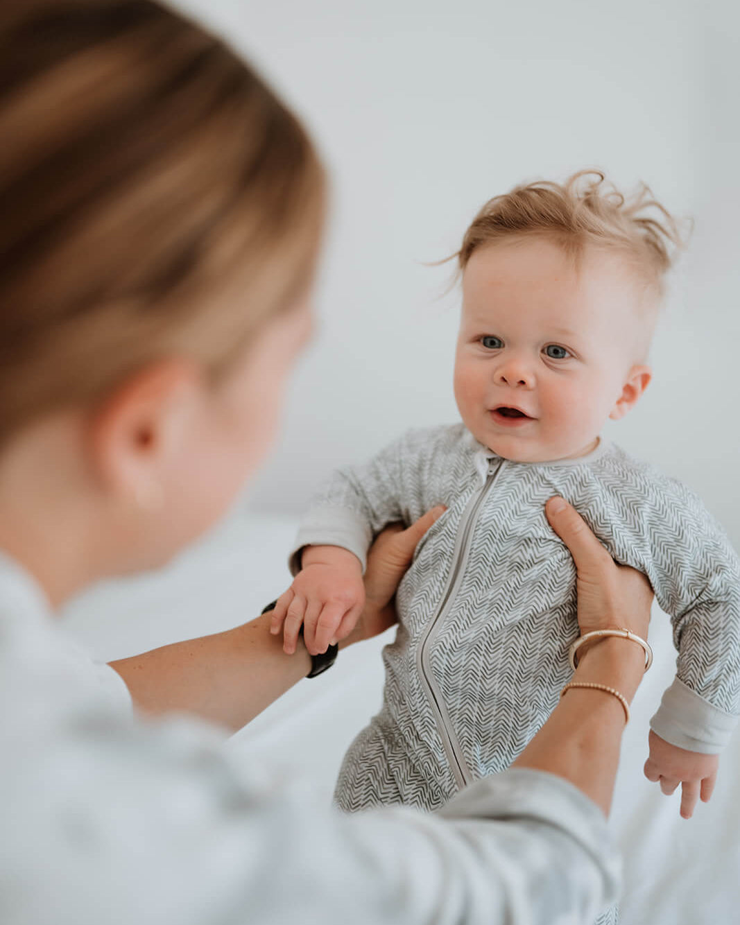 A baby wearing a light gray textured zip-up onesie is being held up by an adult in a softly lit indoor setting.