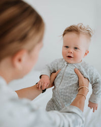 A baby wearing a light gray textured zip-up onesie is being held up by an adult in a softly lit indoor setting.