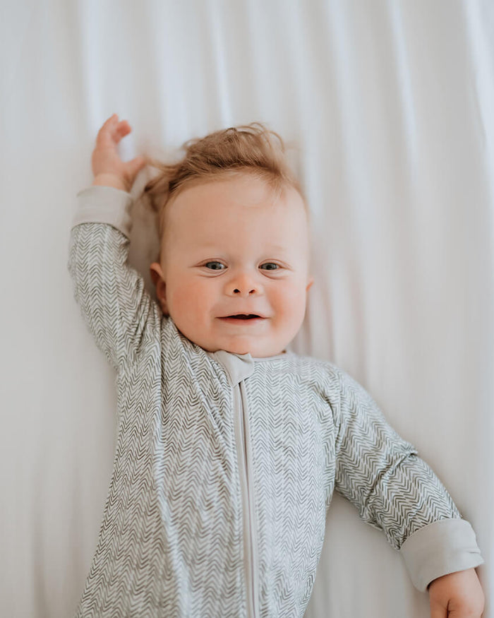 A baby lying on a white bed, wearing a light gray and white textured long-sleeve footless onesie.