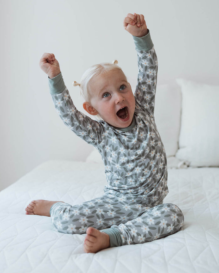 A toddler with blonde hair sitting on a white quilted bed, wearing a long-sleeve pajama set with a gray and white star pattern and raised arms in excitement.