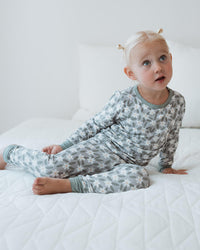 A toddler with blonde hair in two small buns sits on a white quilted bed wearing a long-sleeve pajama set with a gray and white abstract pattern.
