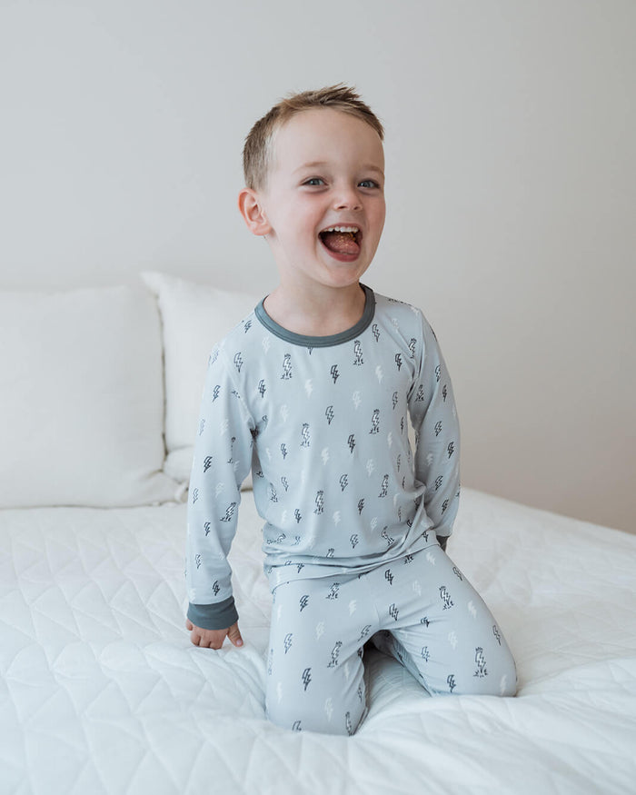 A young boy wearing a light gray long-sleeve pajama set with a small dark pattern, sitting happily on a white quilted bed against a neutral wall.