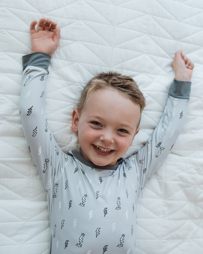 A smiling young boy lies on his back on a white quilted mattress, wearing a light gray long-sleeve pajama with a black tree pattern and dark gray cuffs.