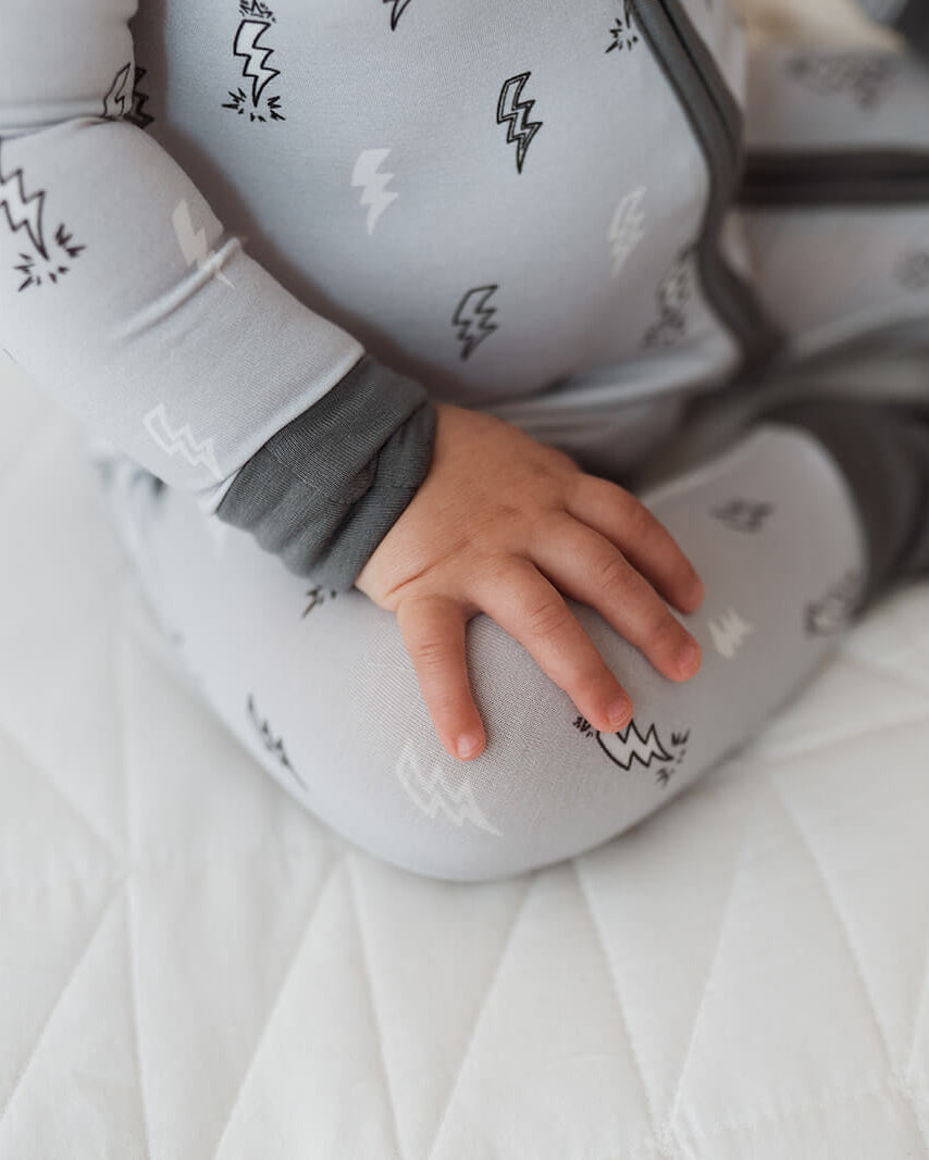 A baby wearing a light gray zip-up sleeper with a black lightning-bolt pattern, sitting on a white quilted mattress.