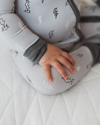 A baby wearing a light gray zip-up sleeper with a black lightning-bolt pattern, sitting on a white quilted mattress.