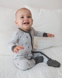 A smiling baby sitting on a white quilted bed wearing a light gray long-sleeve sleeper with dark gray trim and a subtle pattern, paired with dark gray socks.