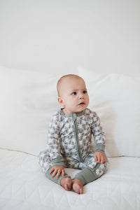 A baby sitting on a white quilted bed wearing a long-sleeve, one-piece zip-up pajama with a gray and white abstract pattern and solid sage green cuffs and collar.