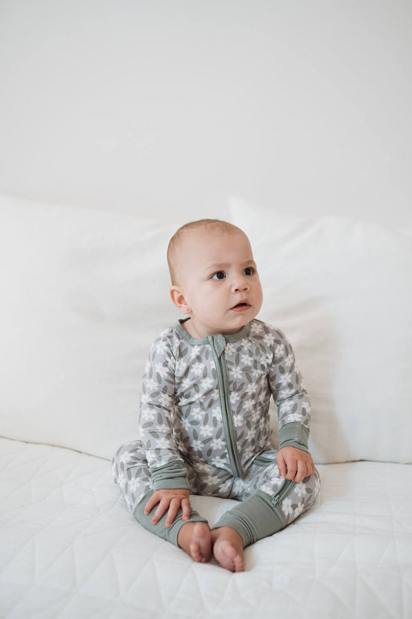 A baby sitting on a white quilted bed wearing a long-sleeve, one-piece zip-up pajama with a gray and white abstract pattern and solid sage green cuffs and collar.