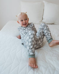 A smiling toddler lying on a white quilted bedspread, wearing a matching long-sleeve pajama set with a gray and white abstract pattern and sage green cuffs.