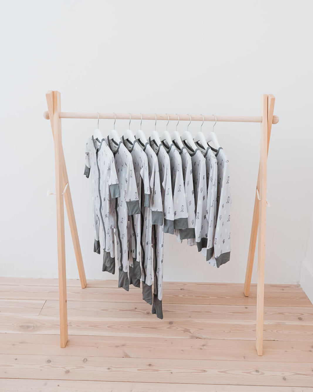 A wooden A-frame clothing rack holding multiple light gray and white patterned baby garments, set against a plain white wall and light wood floor.