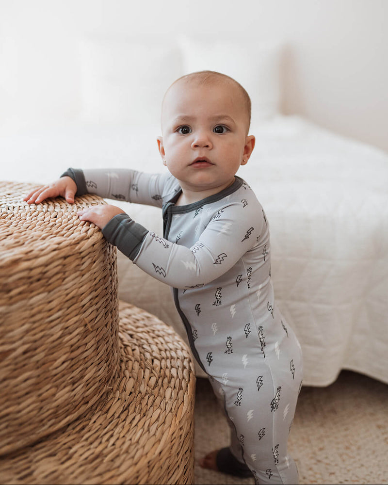 Baby in a gray onesie with animal prints standing next to a woven basket.