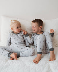 Two young children sitting on a white quilted bed, wearing matching light gray long-sleeve pajamas with a small dark tree print and dark gray cuffs.