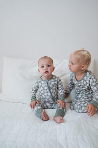 Two toddlers sitting side by side on a white quilted bed, wearing matching long-sleeve pajamas with a gray and white floral pattern and sage green trim.