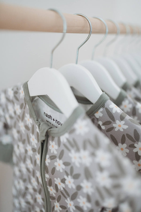 A close-up of baby sleepers hanging on white hangers from a wooden clothing rack. The sleepers feature a soft gray and white floral pattern with a sage green collar trim.