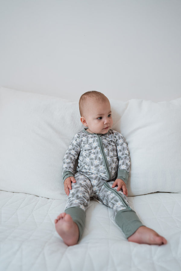 A baby sitting on a white quilted bed wearing a long-sleeve zip-up pajama set with a gray and white animal print and sage green cuffs and hem.